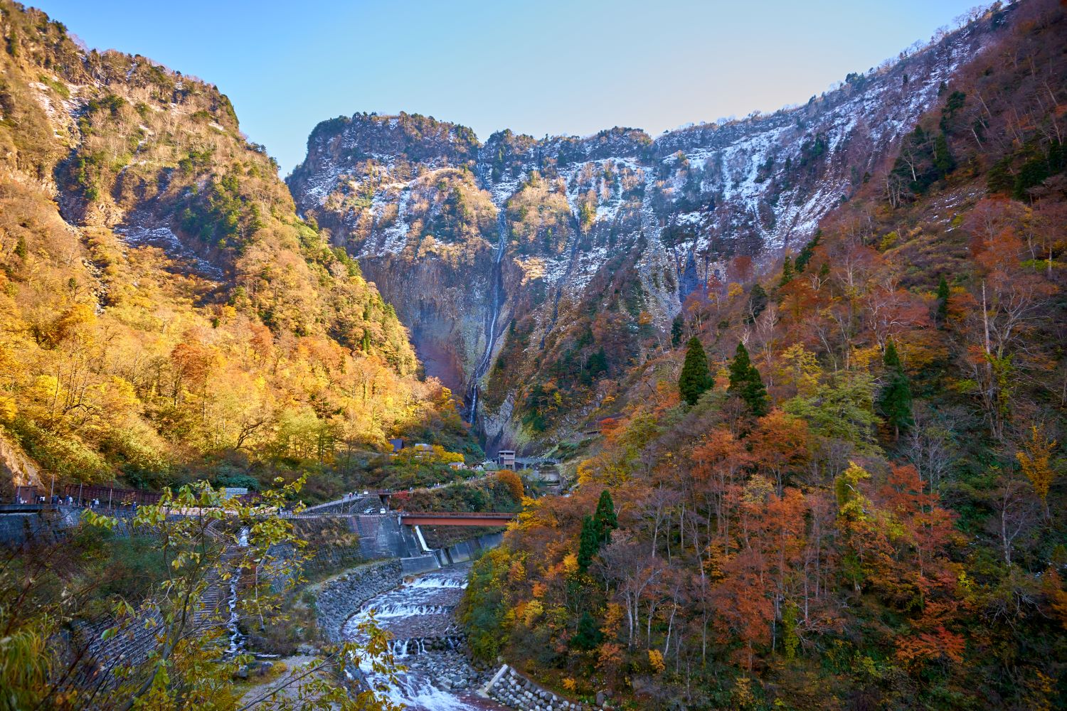 A canyon landscape where autumn foliage on the mountainside and the first snowfall can be seen simultaneously. Shōmyō Falls cascades down in the center background, while a walking path and river spread out in the foreground.