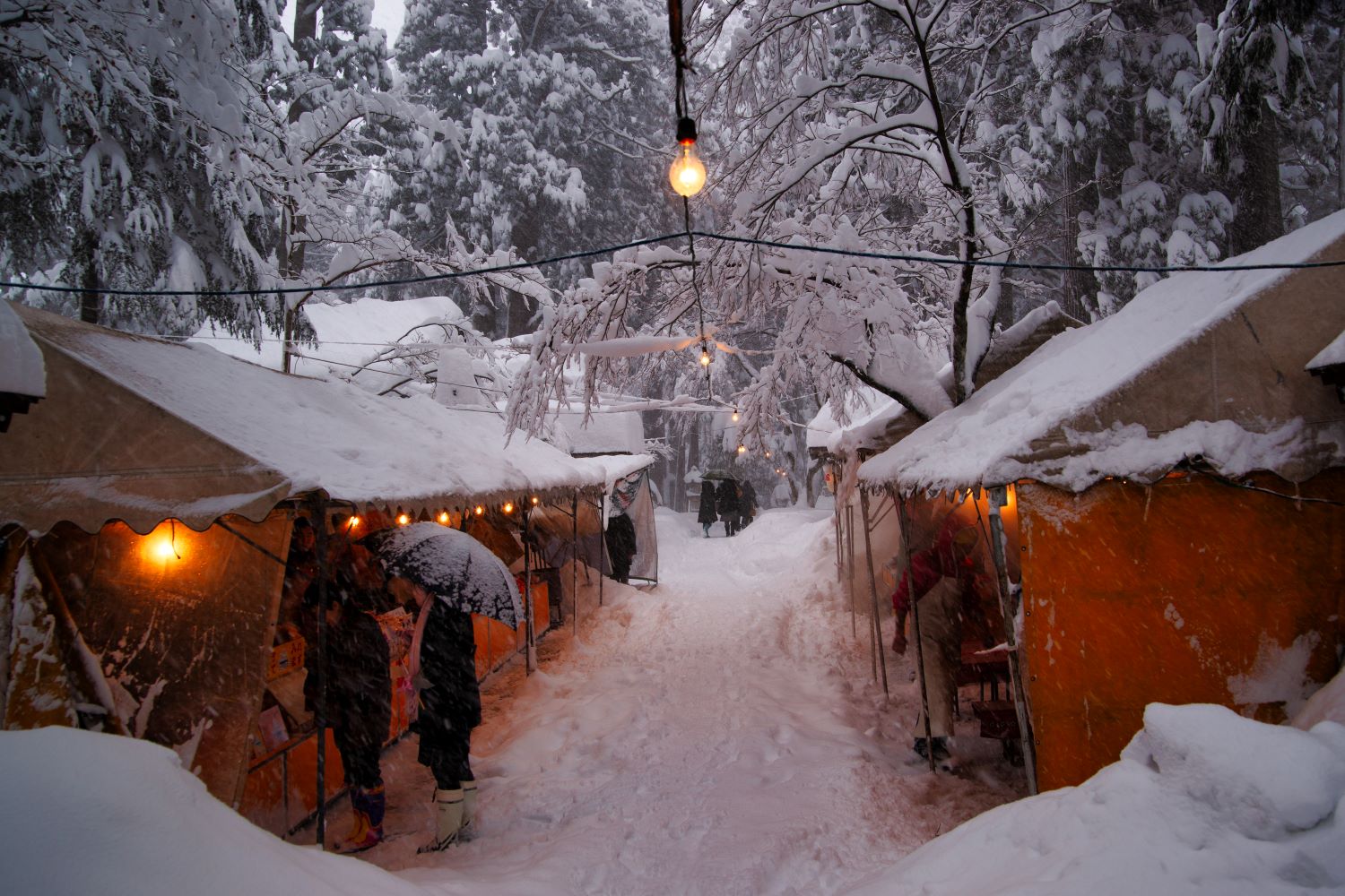 On a snowy winter morning, food stalls line the approach to Oyama Shrine. Orange lanterns illuminate the dimly lit snowy landscape.