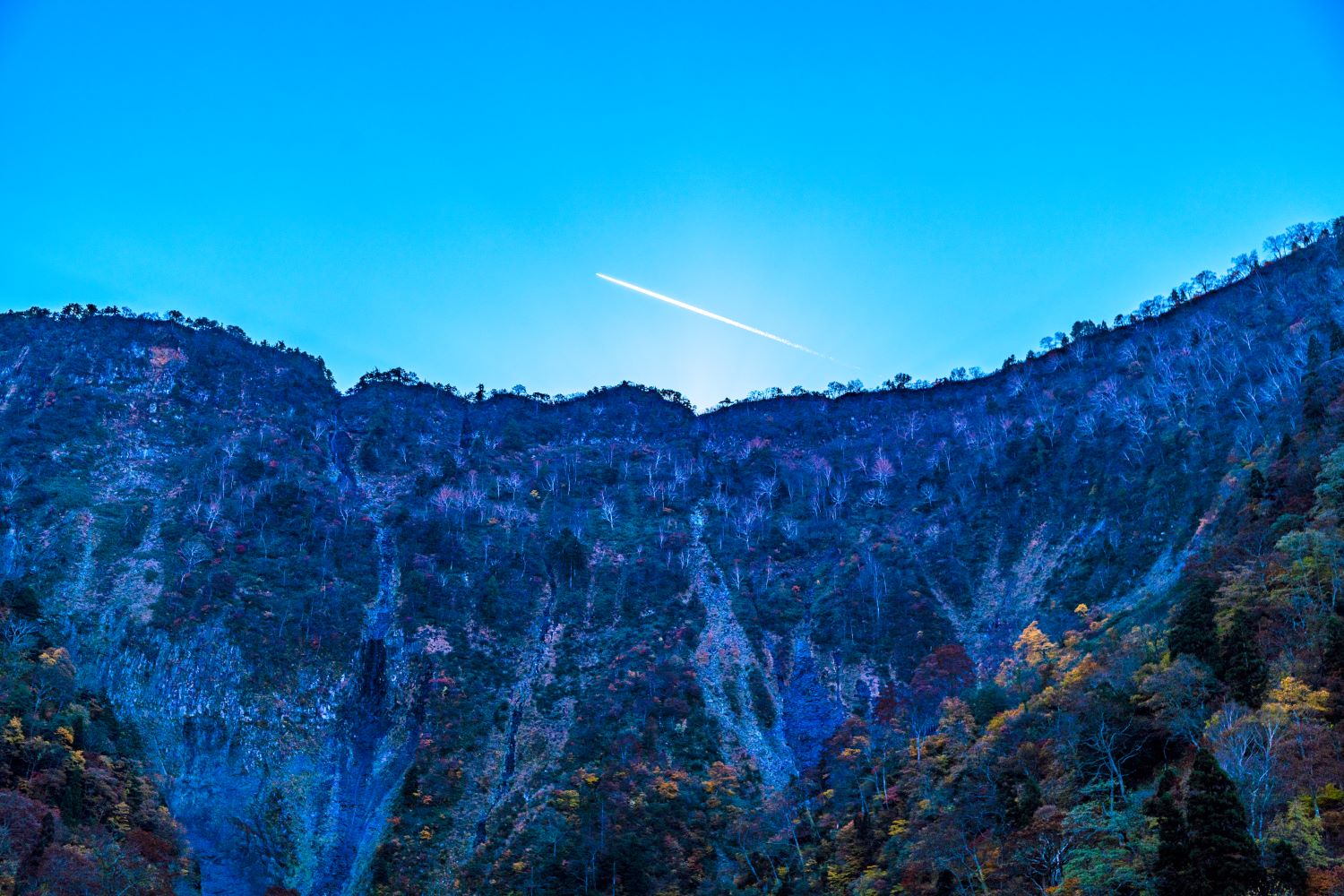 Contrails streak across the blue sky above Shōmyō Falls, mountains shrouded in autumn foliage.