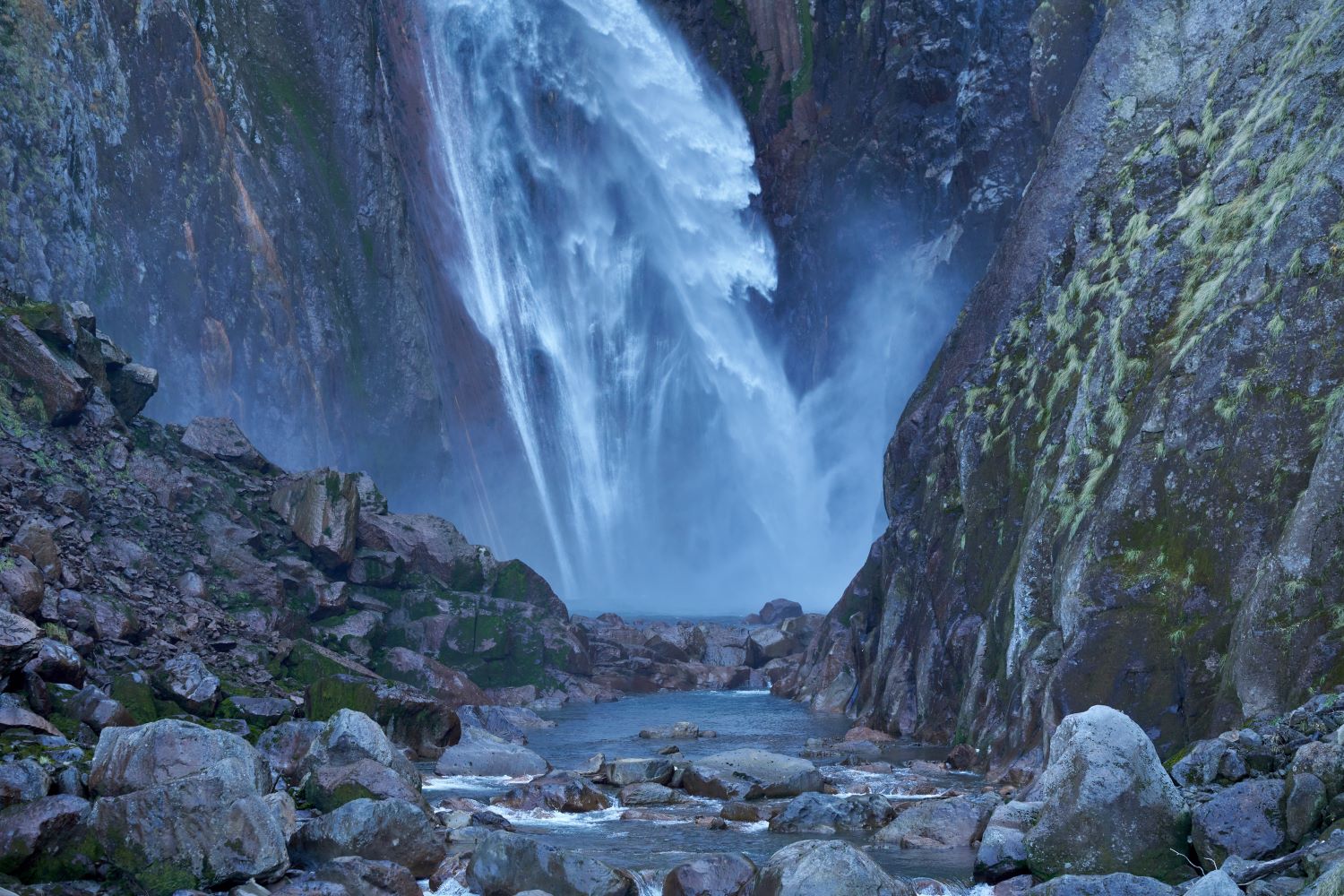 A close-up photograph of the pool at the base of Shōmyō Falls. A massive volume of water cascades down between the rock walls, creating a powerful scene filled with mist.
