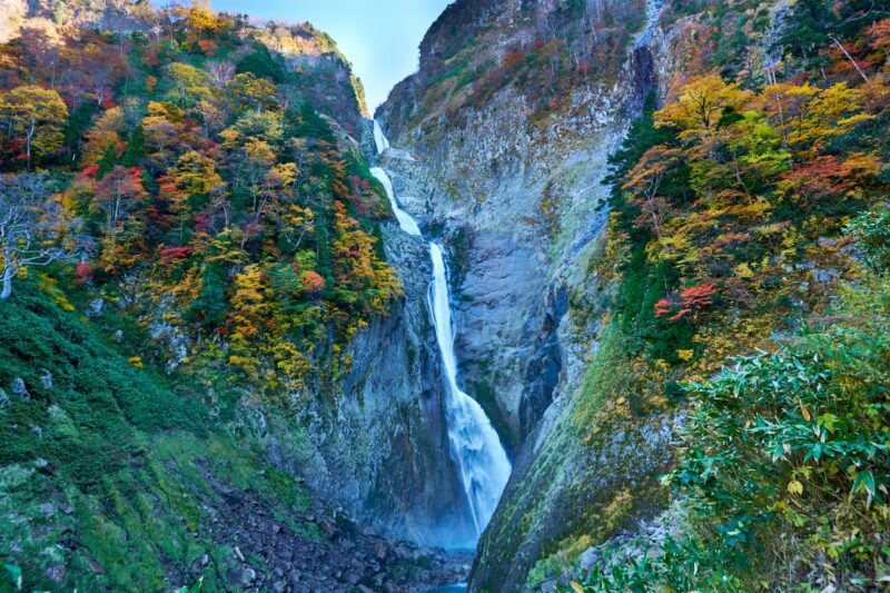 Shōmyō Falls cascading down the cliffs of Mt. Tateyama, adorned with autumn foliage. A stunning autumn vista where the white cascade contrasts vividly with the red and yellow leaves.