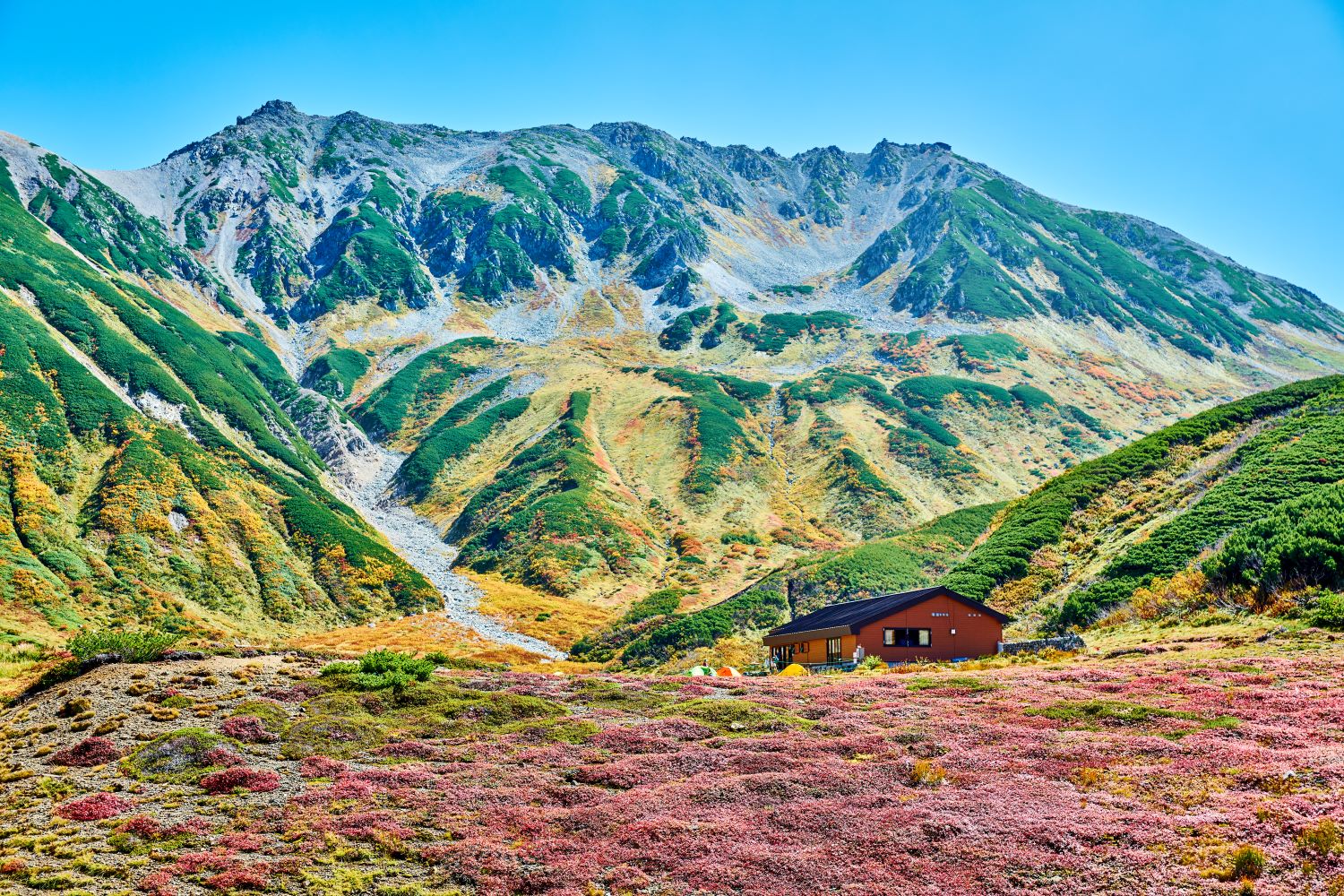 With the mountain hut at Raichōzawa in the foreground, the grasses and leaves turning red and yellow, and the majestic Mt. Tateyama mountain range towering behind.