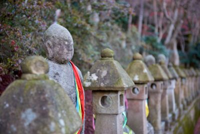 A photo of the approach lined with stone statues of the Gohyaku Rakan and lanterns. Moss-covered stone figures draped in colored cloths, an atmosphere of prayer lingering in the silence.