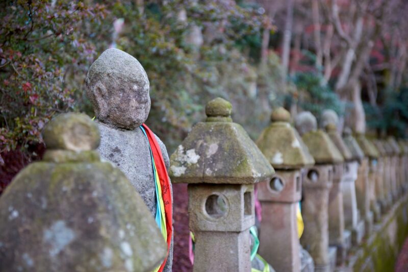 A photo of the approach lined with stone statues of the Gohyaku Rakan and lanterns. Moss-covered stone figures draped in colored cloths, an atmosphere of prayer lingering in the silence.