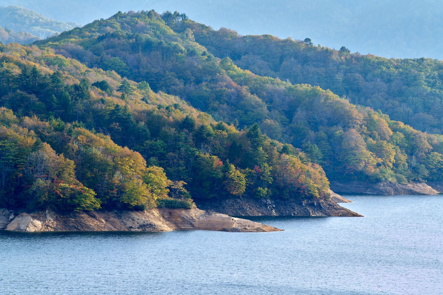 The slopes of the mountains, now ablaze with autumn colors, press close to the shores of Lake Arimine. A landscape unfolds along the lake's surface, where trees dyed red, orange, and yellow spread out.