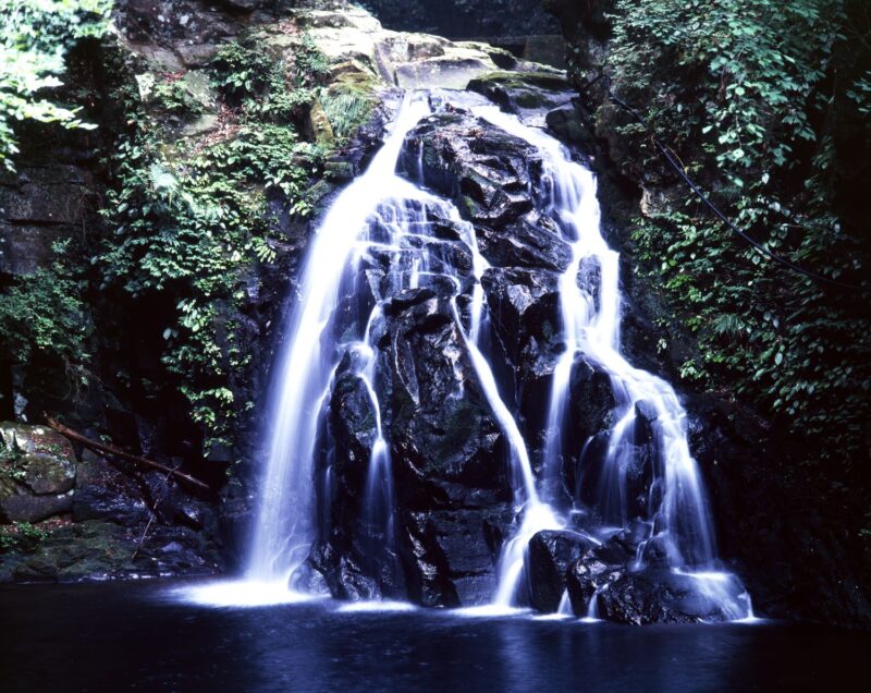 The Akame Forty-Eight Waterfalls and Senju Falls cascade down the rock face in multiple streams, white as snow. They are surrounded by black rock surfaces and green moss.