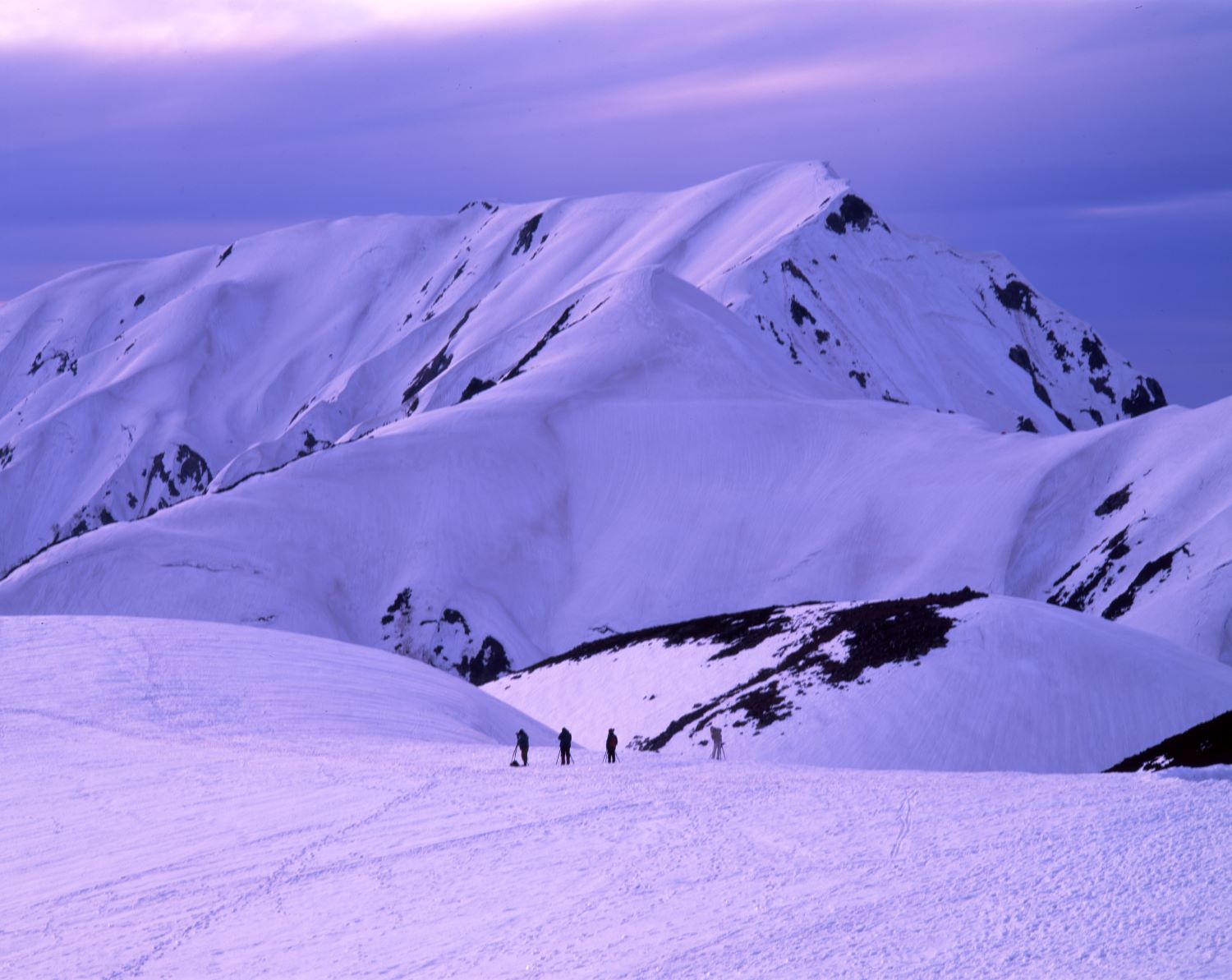 Mountaineers climbing the snow-covered ridges of Mount Oku-Dainichi. An early winter evening scene tinged with purple light.
