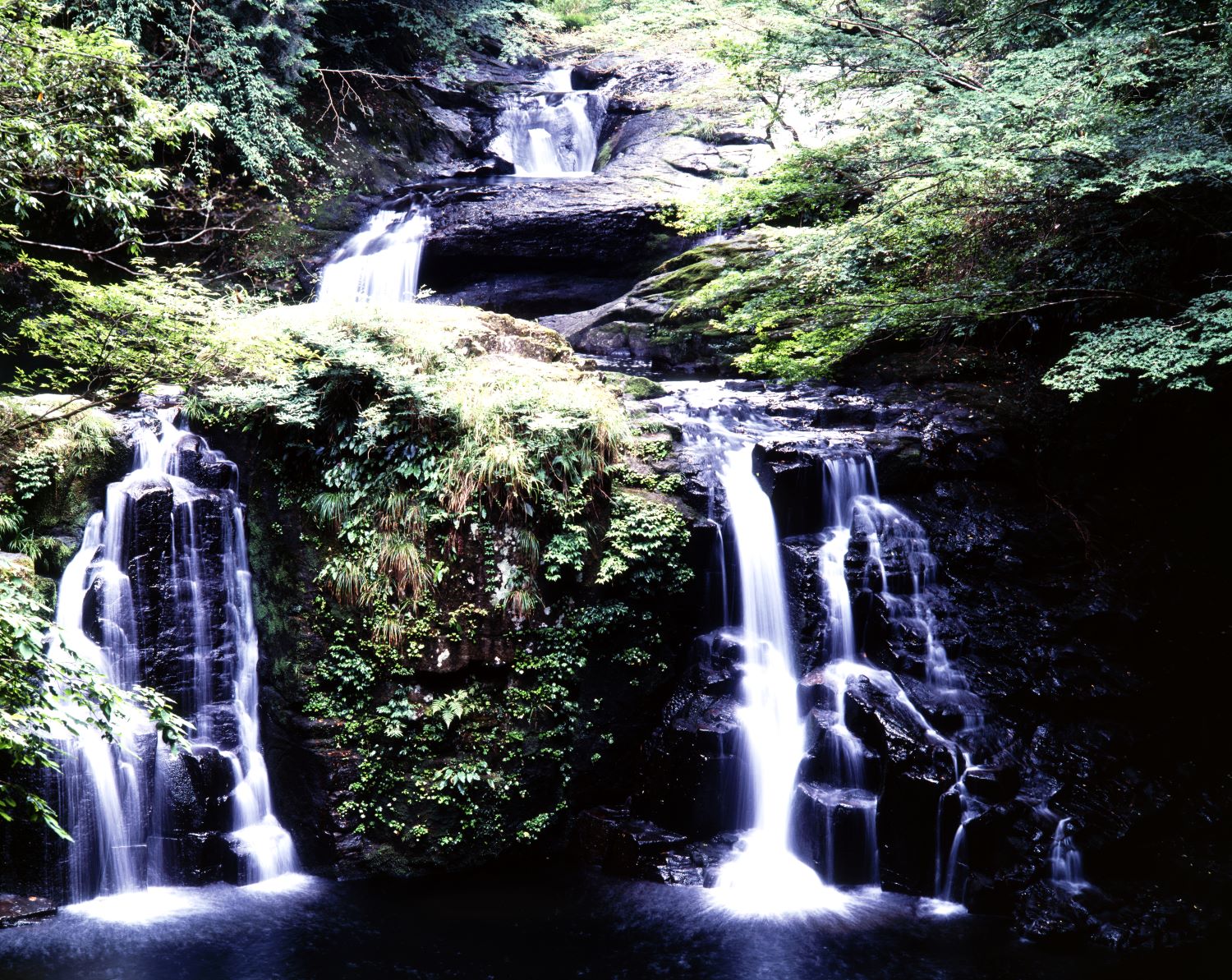 Akame Forty-Eight Waterfalls and Nunobiki Falls cascade down the approximately 30-meter-high rock face, splitting into multiple streams that flow like white cloth draped down. The surrounding area is covered in greenery.