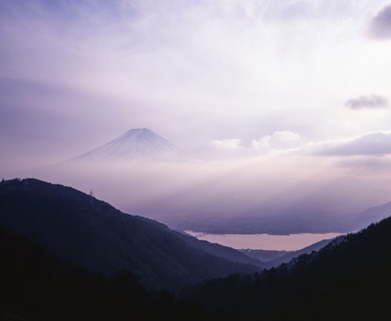 A landscape photograph showing Mount Fuji enveloped in mist and soft light, with Lake Kawaguchi spreading out at its base.