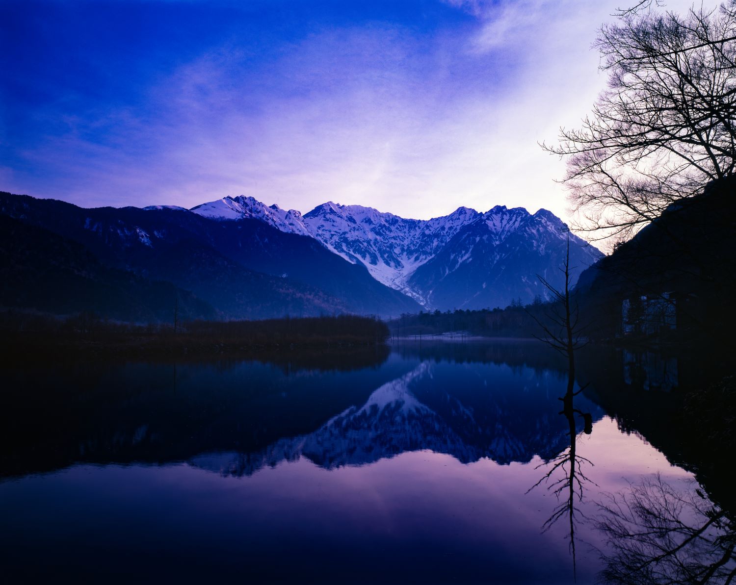 The Hotaka Range viewed from Taisho Pond in Kamikochi. A landscape photograph showing the mountain ridges, crowned with their first snow, reflected on the lake's surface.