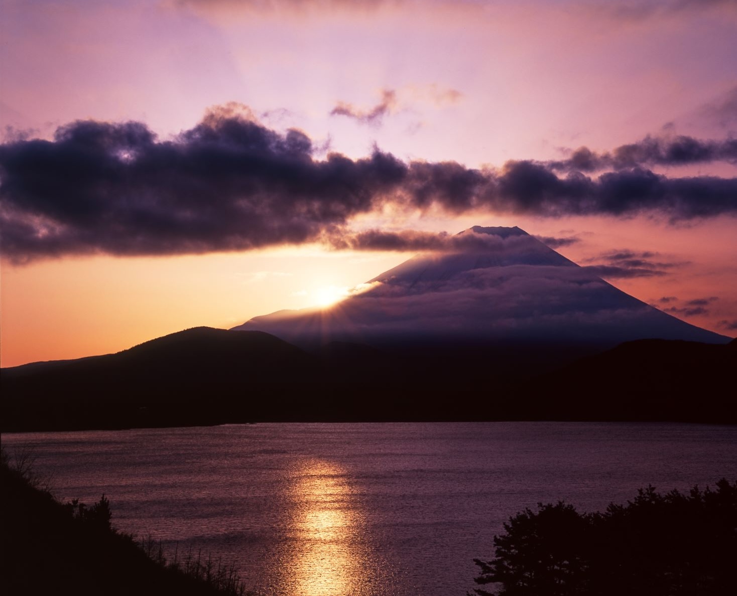 A landscape photograph showing Mount Fuji shrouded in clouds and the rising sun reflected on the surface of Lake Motosu at dawn.