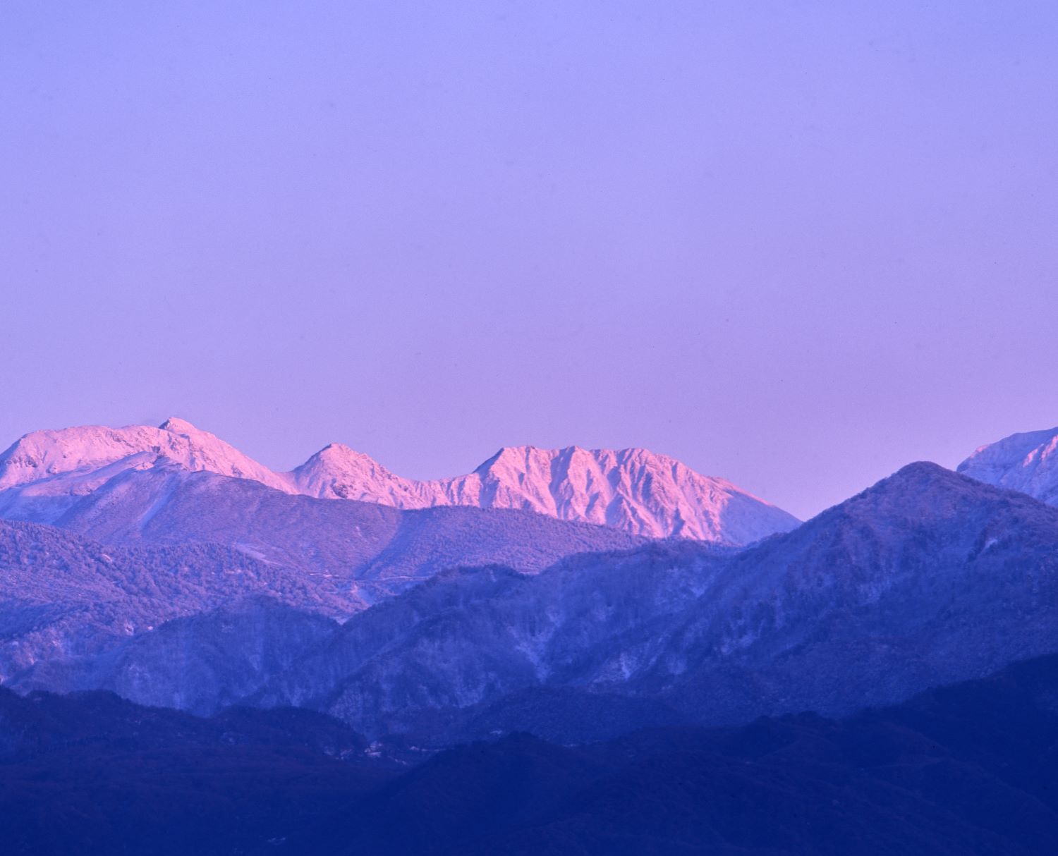 A mountain photograph capturing Mt. Oyama of the Mt. Tateyama Range bathed in the light of dusk from a distant viewpoint. Multiple mountain ridges spread out in the foreground.