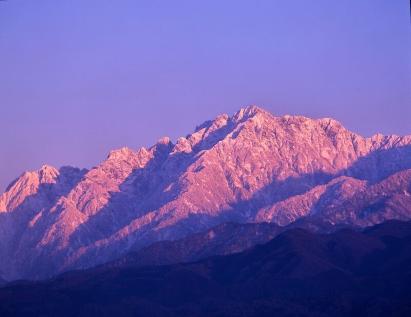 In the evening's oblique light, Mount Tsurugi, cloaked in snow, glows crimson-purple in the distance.