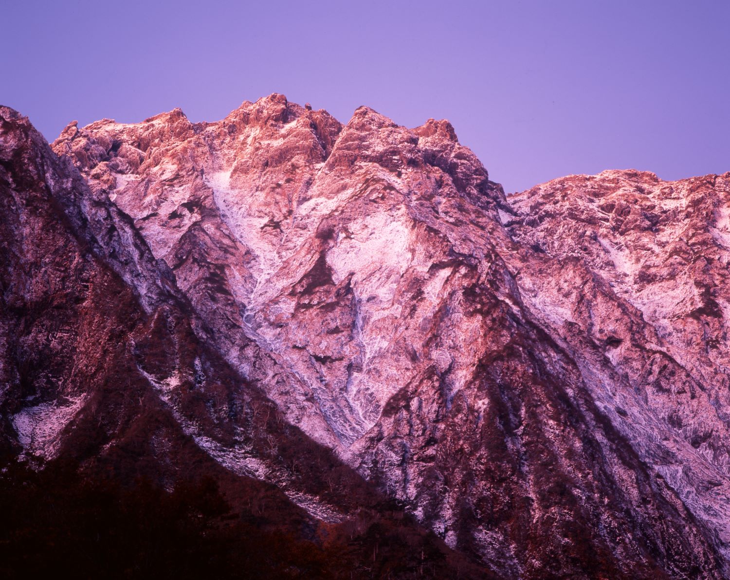 A close-up photograph of the rock face of Tanigawa-dake's Ichinokurasawa, bathed in the morning light and glowing red against a pale purple sky.