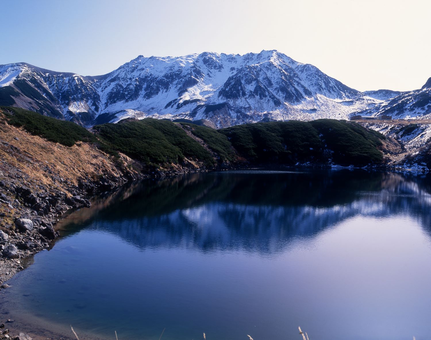 The snow-capped Mt. Tateyama mountain range forms the backdrop, while the winter landscape reflects the mountain ridges upon the surface of Mikurigaike.