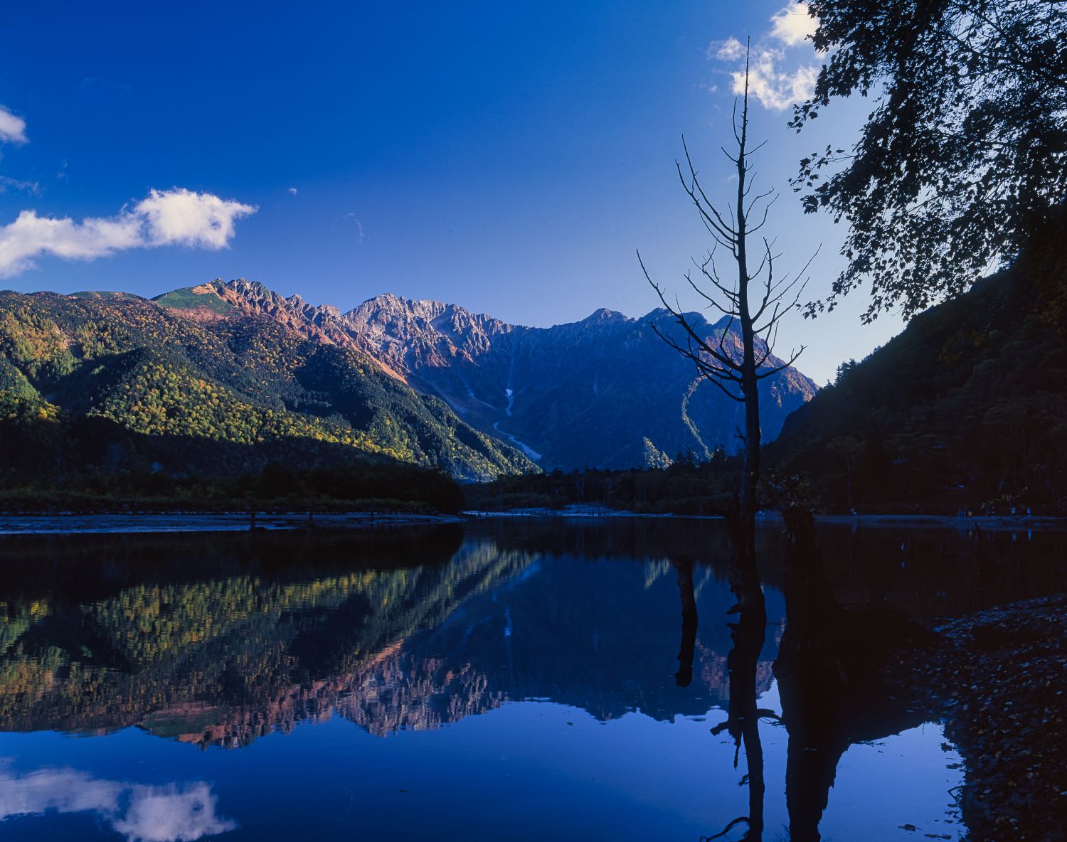 The Hotaka Range viewed from Lake Taisho in autumn. A landscape photograph capturing the reflection of the rock faces and mountain silhouettes, now ablaze with autumn colors, on the lake's surface.