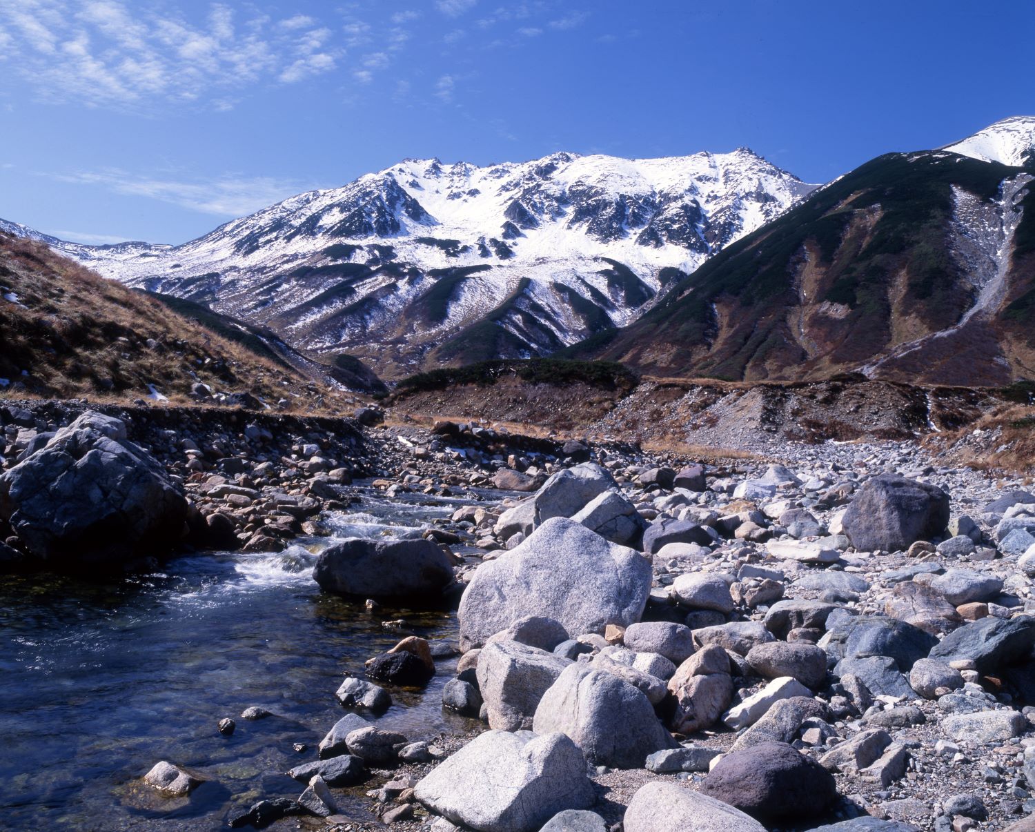 Beyond the clear stream and rocky outcrops of Raichōzawa, the snow-capped Mt. Tateyama mountain range spreads out in the early winter landscape.