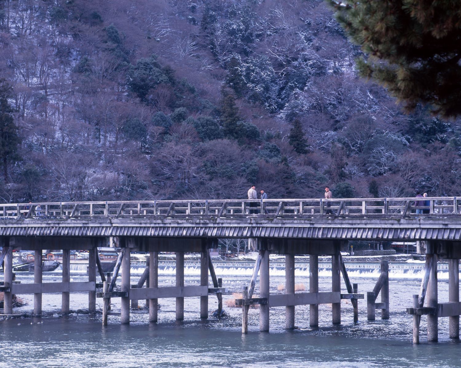 Amidst the snow lingering on the mountainside of Arashiyama, people walk across the Togetsukyo Bridge spanning the Oi River.