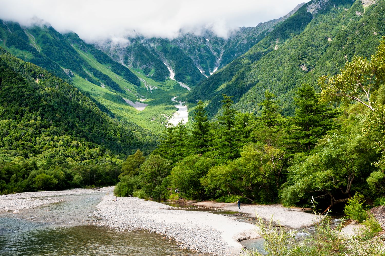 A view of the Hotaka mountain range from the riverbank of the Azusa River in Kamikochi. The lush green forest and the white snowfields lingering in the valley spread out impressively.