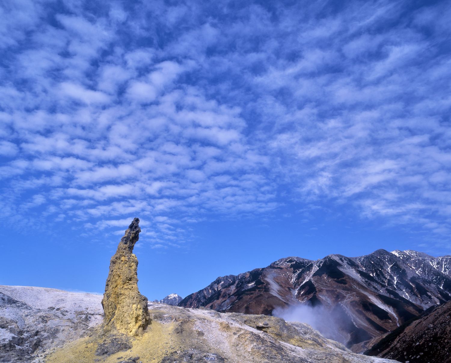The sulfur-stained rock towers of Mt. Tateyama Jigokudani and the Tateyama mountain range towering behind it, clouds floating against the blue sky.