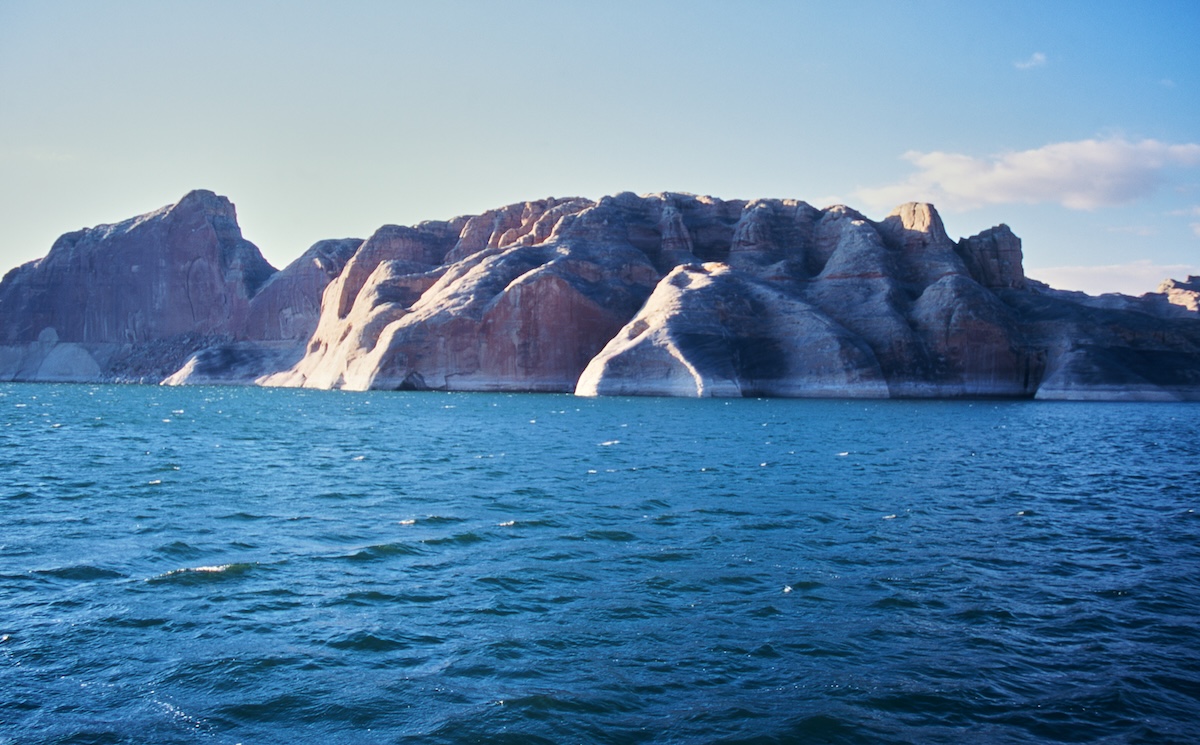 The reddish-brown rock walls of Lake Powell and the blue lake surface illuminated by the light before dusk