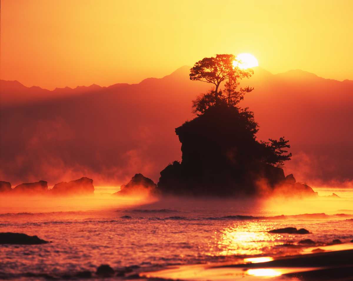 Sunrise at Amaharashi Coast with sea smoke, sunlight emerging beside Onna-iwa, and Mt. Tsurugi glowing faintly in the background.